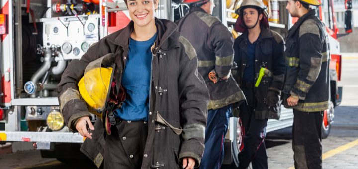 Portrait of happy firewoman with male colleagues discussing by truck in background at fire station