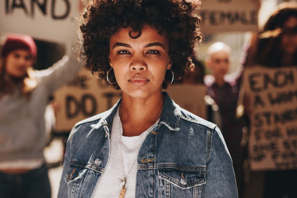 Young african woman standing outdoors with group of demonstrator in background. Woman protesting with group of activists outdoors on road.