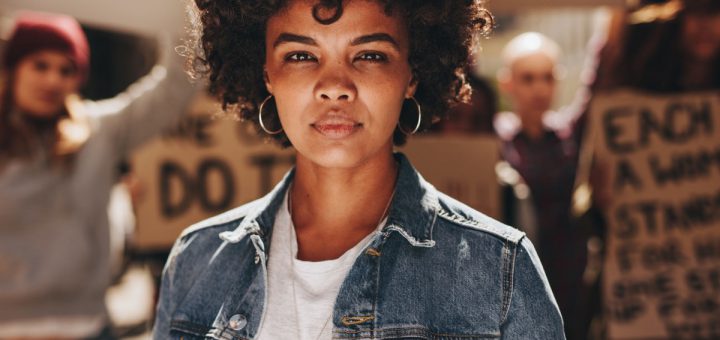 Young african woman standing outdoors with group of demonstrator in background. Woman protesting with group of activists outdoors on road.
