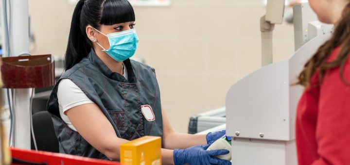 A young woman in a medical mask and gloves, working at the checkout in a supermarket. In the foreground is a customer in a blur. Close up. Concept of coronovirus, protection from infection and industrial crisis.