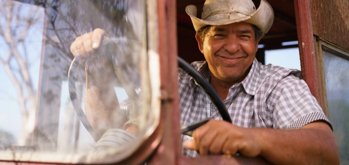 Farming and cultivations in Latin America. Portrait of middle aged hispanic farmer sitting proud in his tractor at sunset, holding the steering wheel. He looks at the camera and smiles happy.