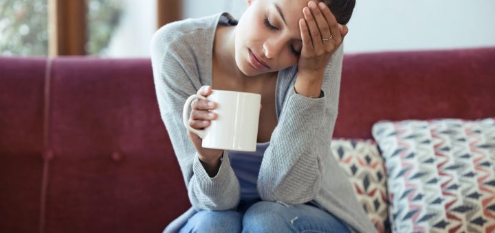 Shot of depressed young woman having headache while drinking coffee on sofa at home.