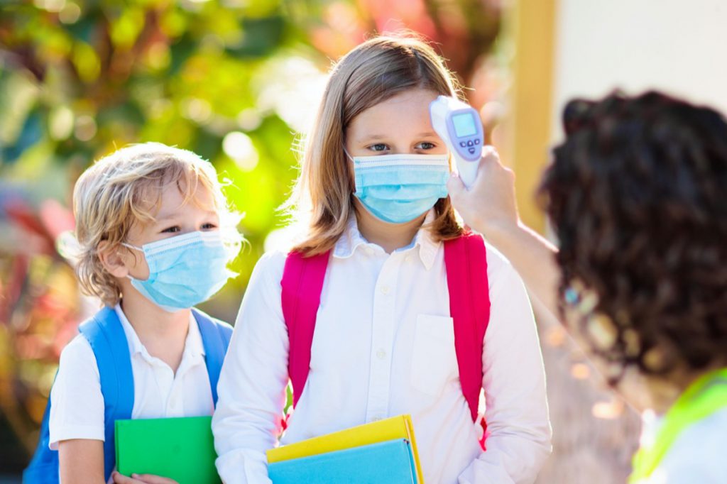 Temperature screening and medical check at school. Child in face mask in class in covid-19 outbreak. Teacher with thermometer at preschool entrance. Social distancing. Coronavirus prevention.