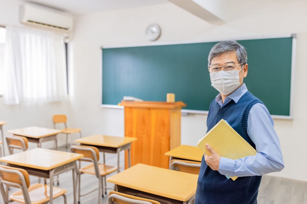 asian senior male professor wearing face mask hold books and look at you in front of blackboard at college classroom