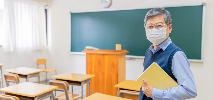 asian senior male professor wearing face mask hold books and look at you in front of blackboard at college classroom