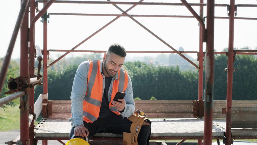 Person working in construction site. White young man at work in new house, smiling and using smartphone for internet and social media during break