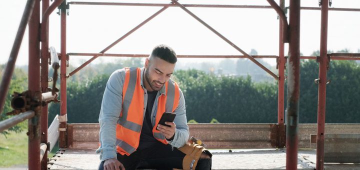 Person working in construction site. White young man at work in new house, smiling and using smartphone for internet and social media during break