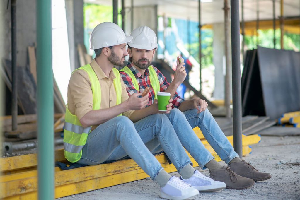Building site. Building workers in yellow vests and helmets sitting on boards, having coffee with sandwiches, discussing something