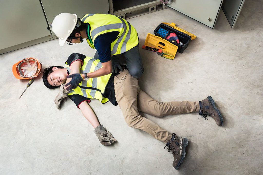 A worker helps a fellow worker who has fallen, both wear hardhats and yellow vests; Workers’ Compensation concept