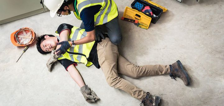 A worker helps a fellow worker who has fallen, both wear hardhats and yellow vests; Workers’ Compensation concept