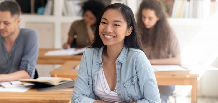 Smiling smart female asian teen girl looking at camera sitting at desk studying in classroom with diverse classmates group, happy chinese college university student in international class portrait