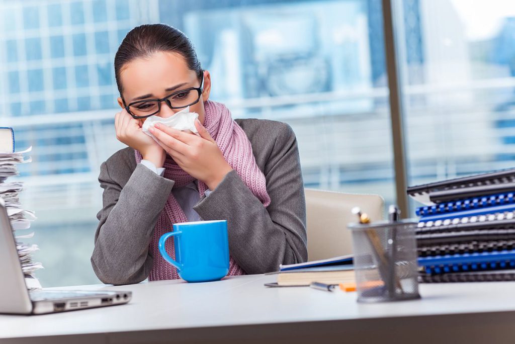 Professional woman at desk, holding a tissue to her nose and leaning on her hand; paid time off concept