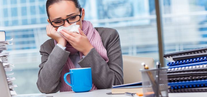 Professional woman at desk, holding a tissue to her nose and leaning on her hand; paid time off concept