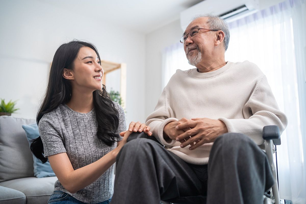 Young woman kneeling next to elderly man in wheelchair; family benefits concept