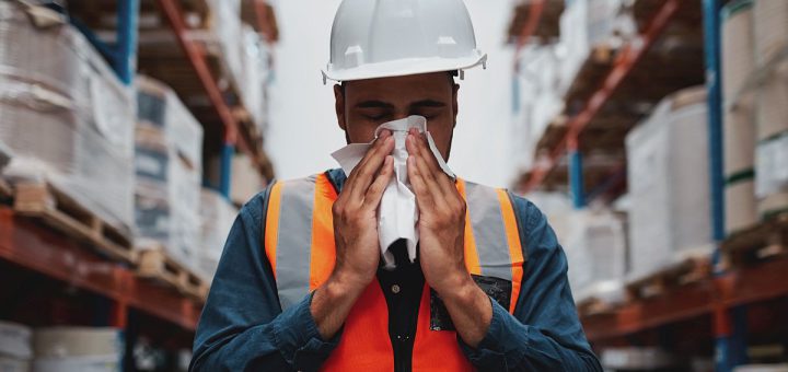 Worker wearing a hardhat in a warehouse blowing his nose; paid sick leave concept