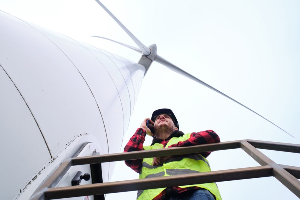 View from below of engineer wearing hard hat and reflective vest talking on mobile phone on wind turbine stairway; inflation reduction act benefits unions concept