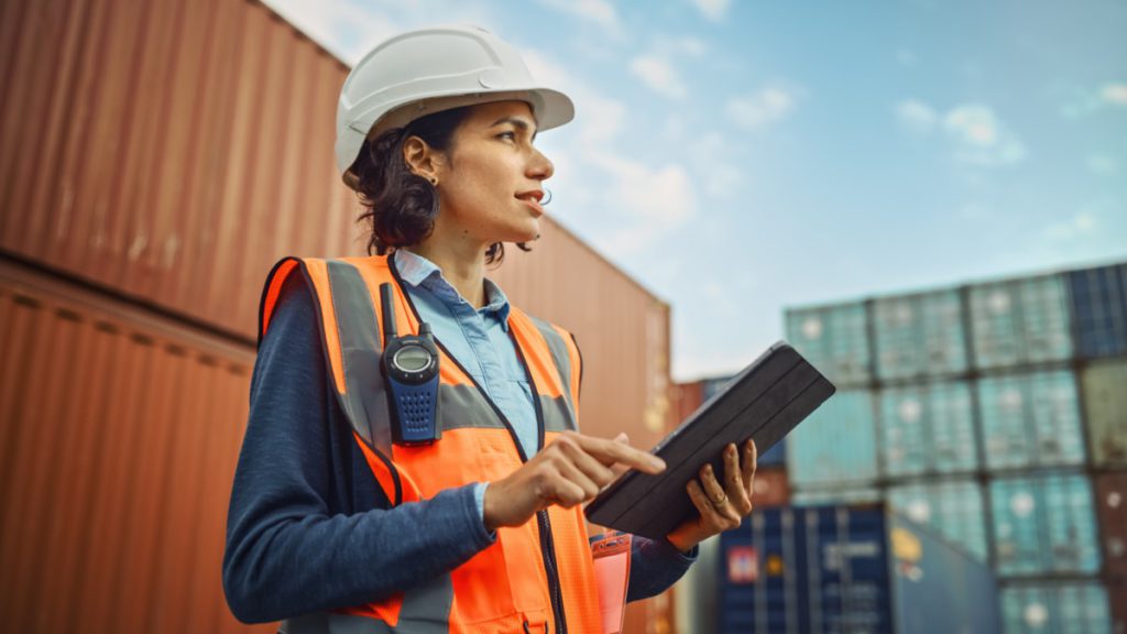Worker in white hard hat and high-visibility vest working on tablet in container terminal; Department of Labor initiatives concept