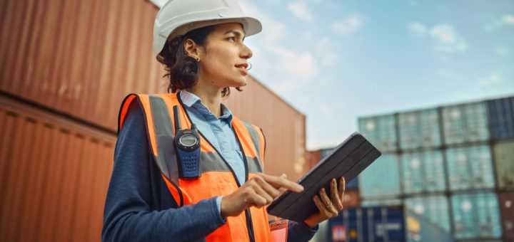 Worker in white hard hat and high-visibility vest working on tablet in container terminal; Department of Labor initiatives concept